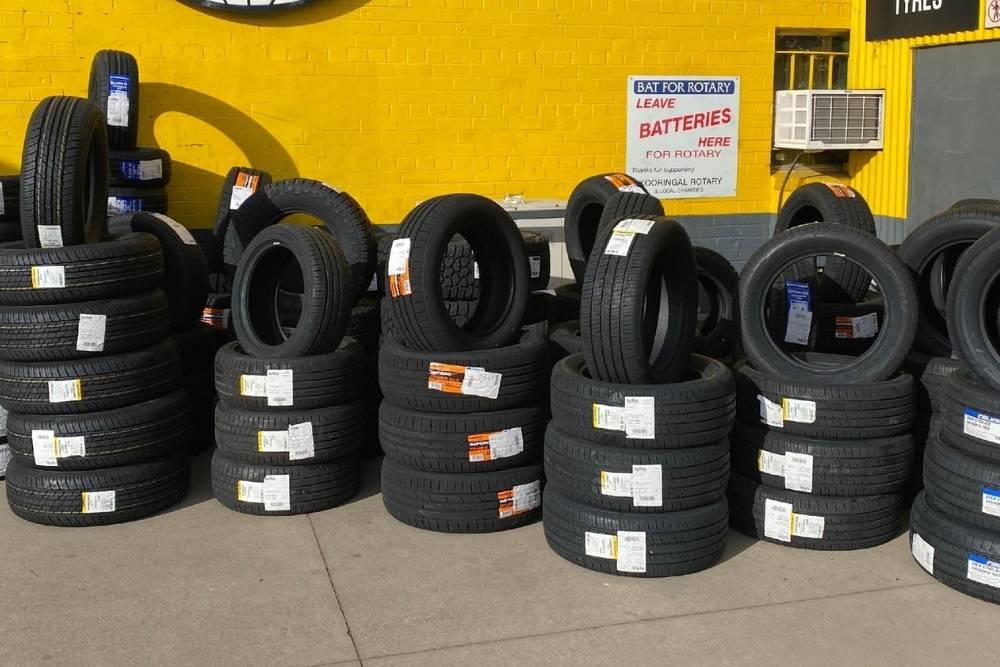 Stacks of black vehicle tyres with white stickers displayed outside against a bright yellow brick wall-